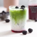 Iced matcha latte with three layers of matcha, milk, and blackberry syrup in a tall glass garnished with blackberries.