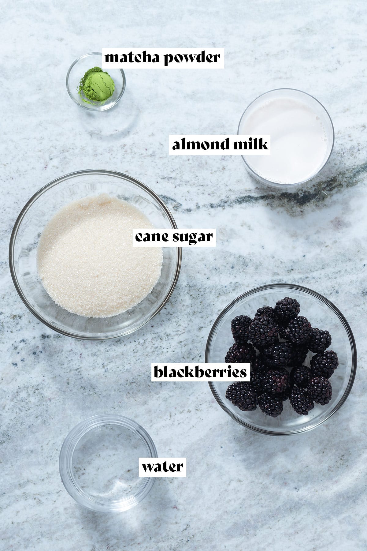 Blackberries, cane sugar, milk, matcha powder, and water laid out on a grey stone background.