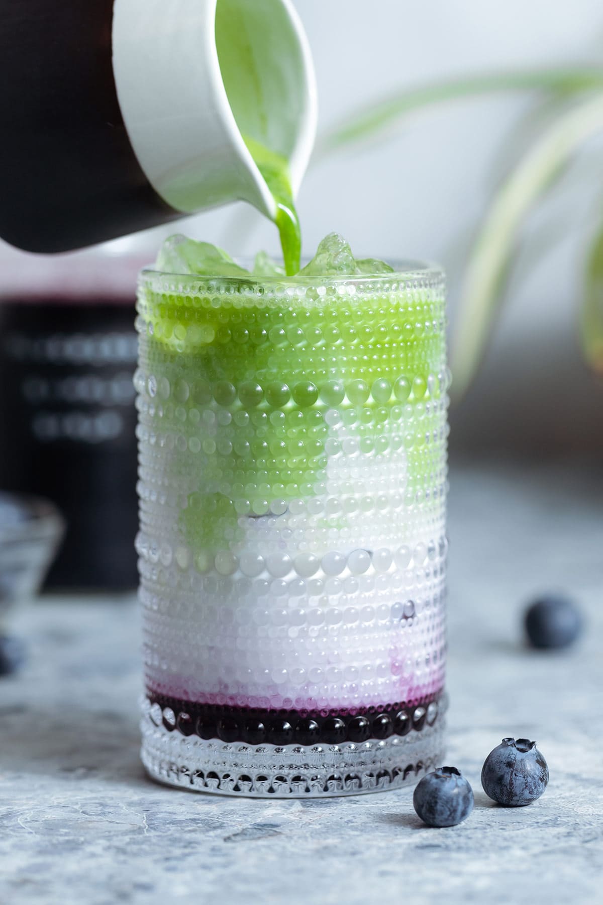 Matcha being poured over milk and blueberry syrup into a tall glass over ice.