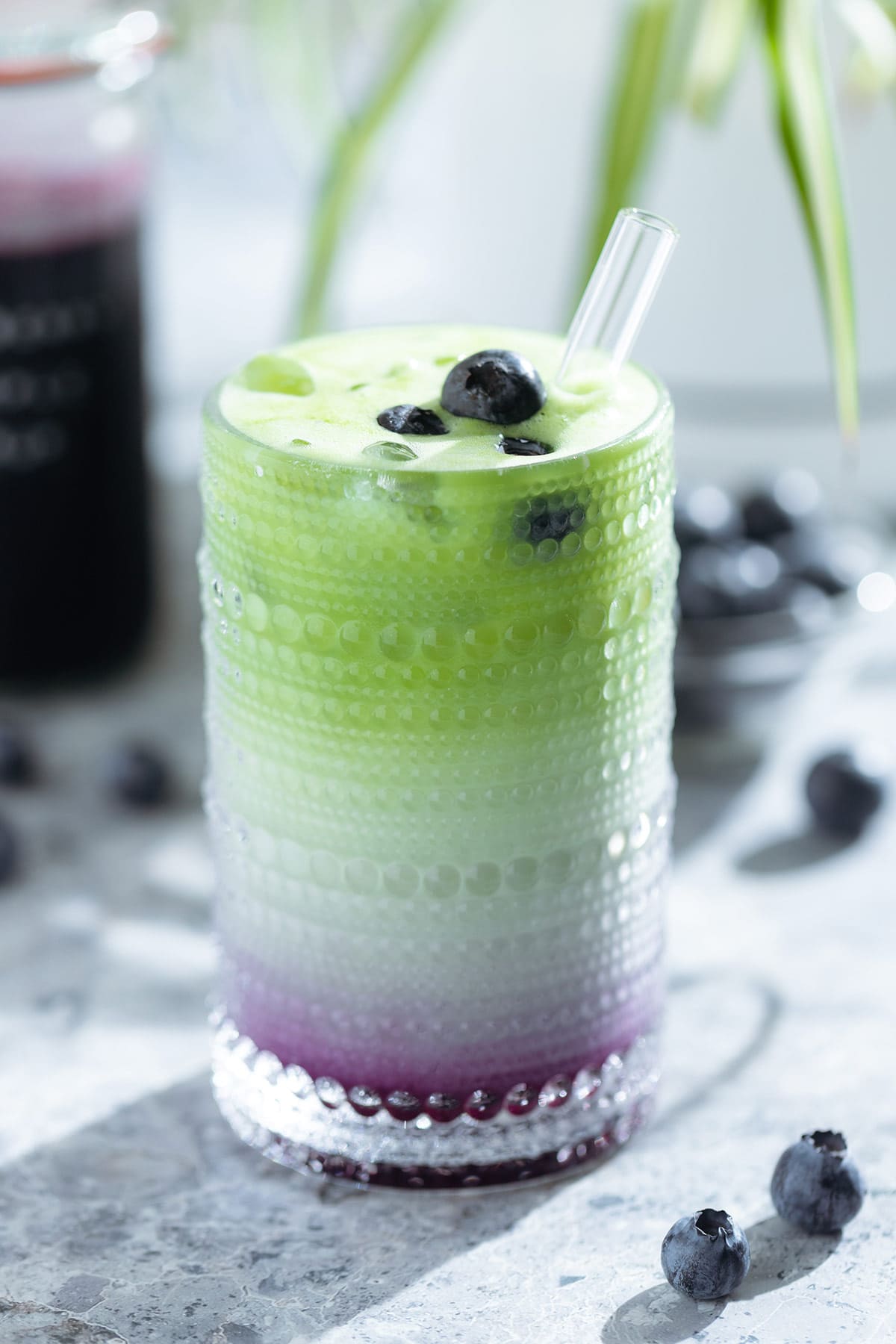 Iced matcha latte in a tall glass with a layer of blueberry syrup on the bottom of the glass garnished with fresh blueberries with a glass straw and a bottle of blueberry syrup in the background.