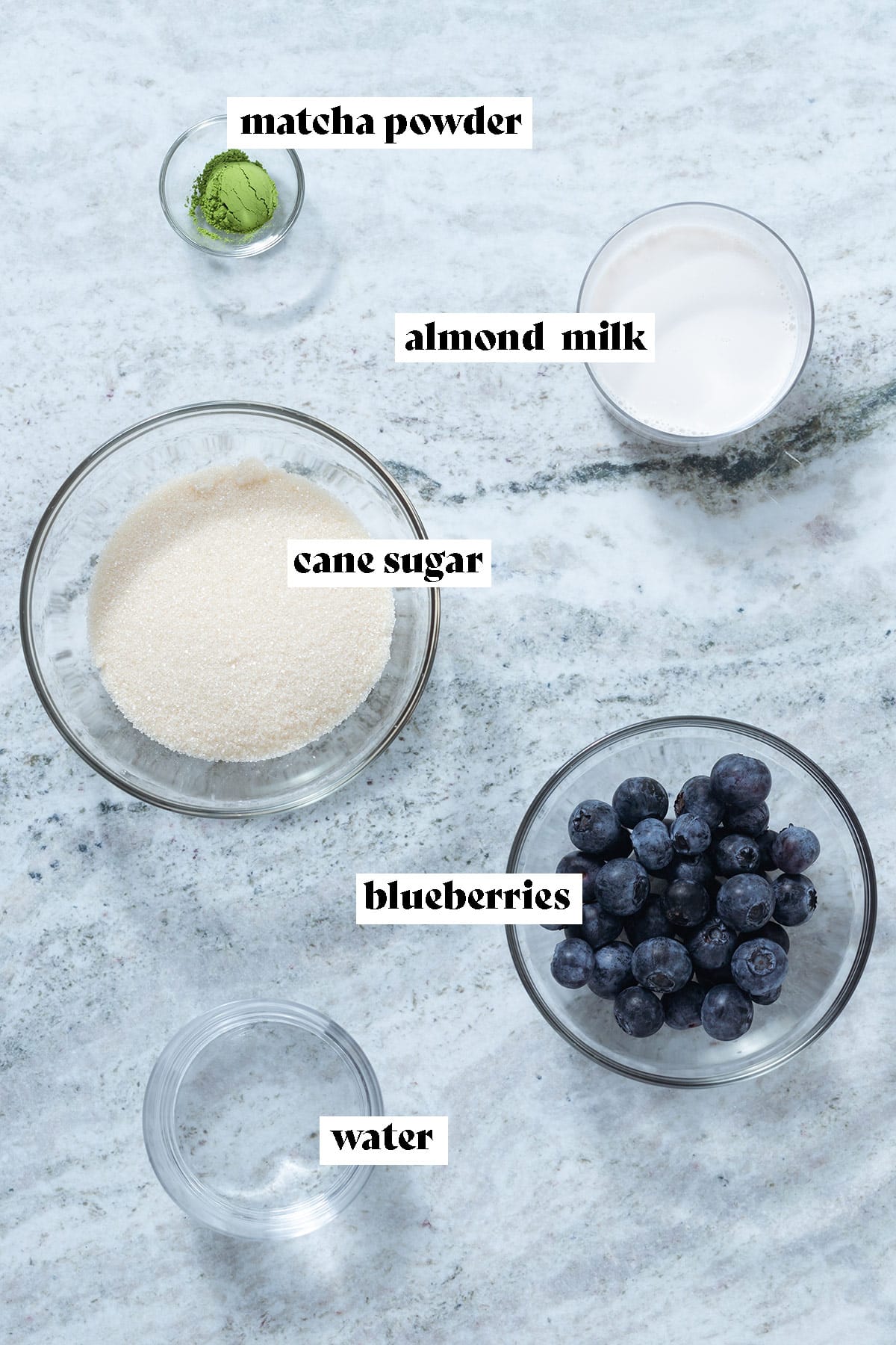 Blueberries, cane sugar, matcha powder, water, and almond milk laid out on a grey stone background.