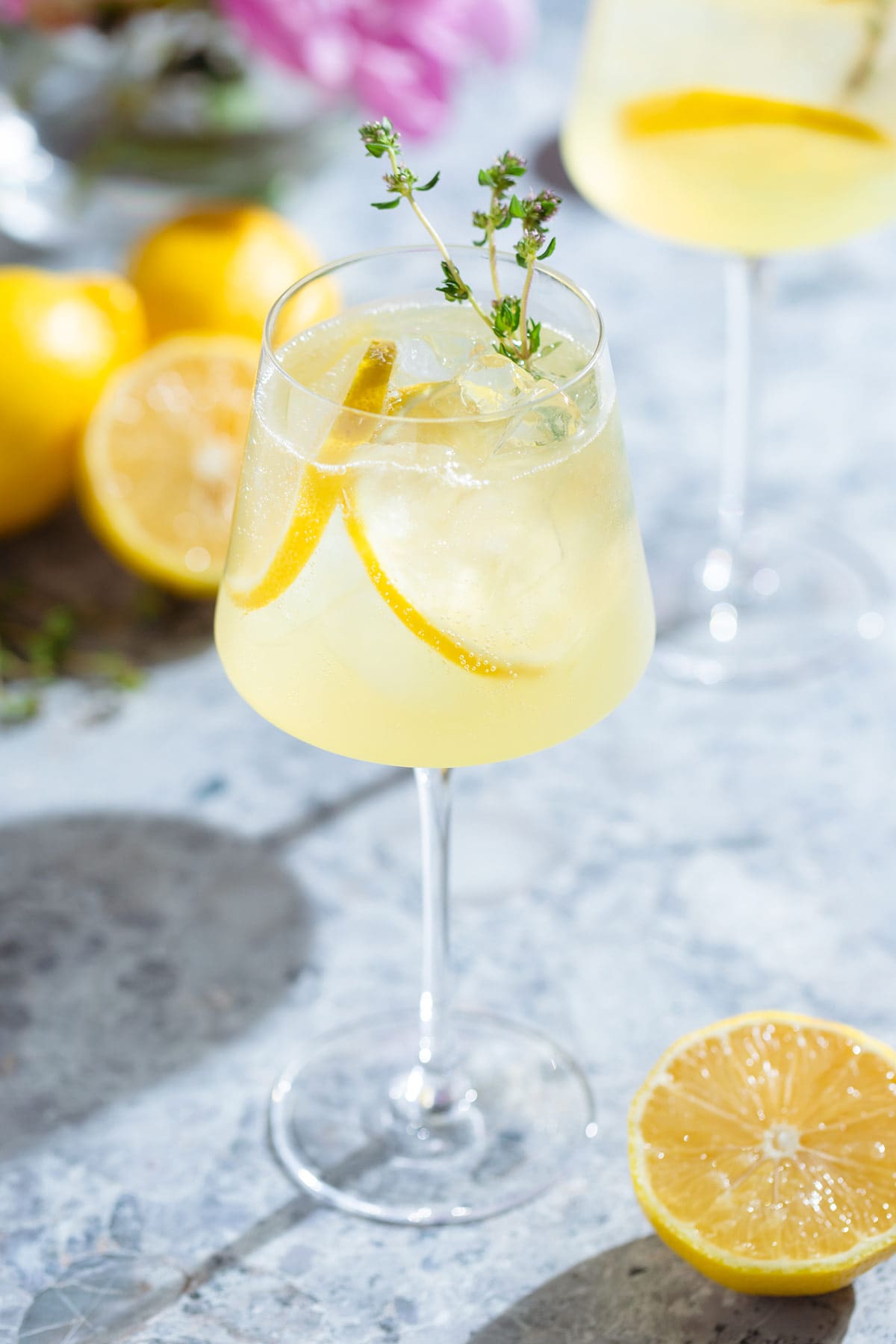 A tall white wine glass with a lemon spritzer garnished with lemon slices and fresh thyme with more lemons around the glass.