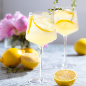 A tall white wine glass with a lemon spritzer garnished with lemon slices and fresh thyme with more lemons around the glass.