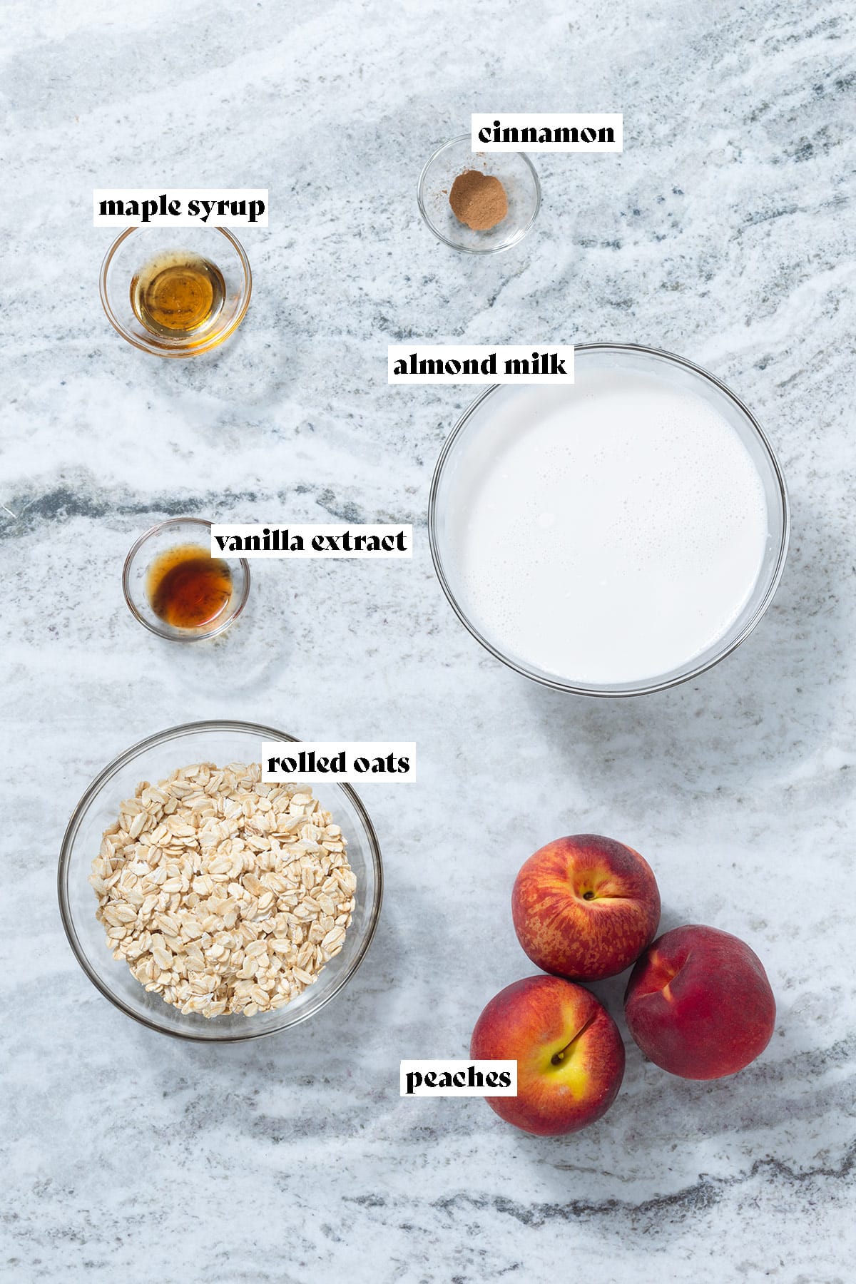 Oats, almond milk, peaches, maple syrup and other ingredients laid out on a grey stone background.
