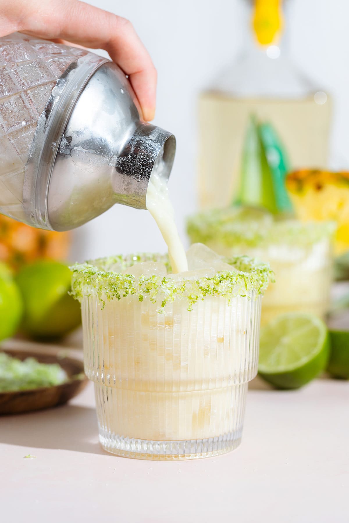 Creamy pineapple margarita being poured into a short glass with ice and lime zest and salt around rim.