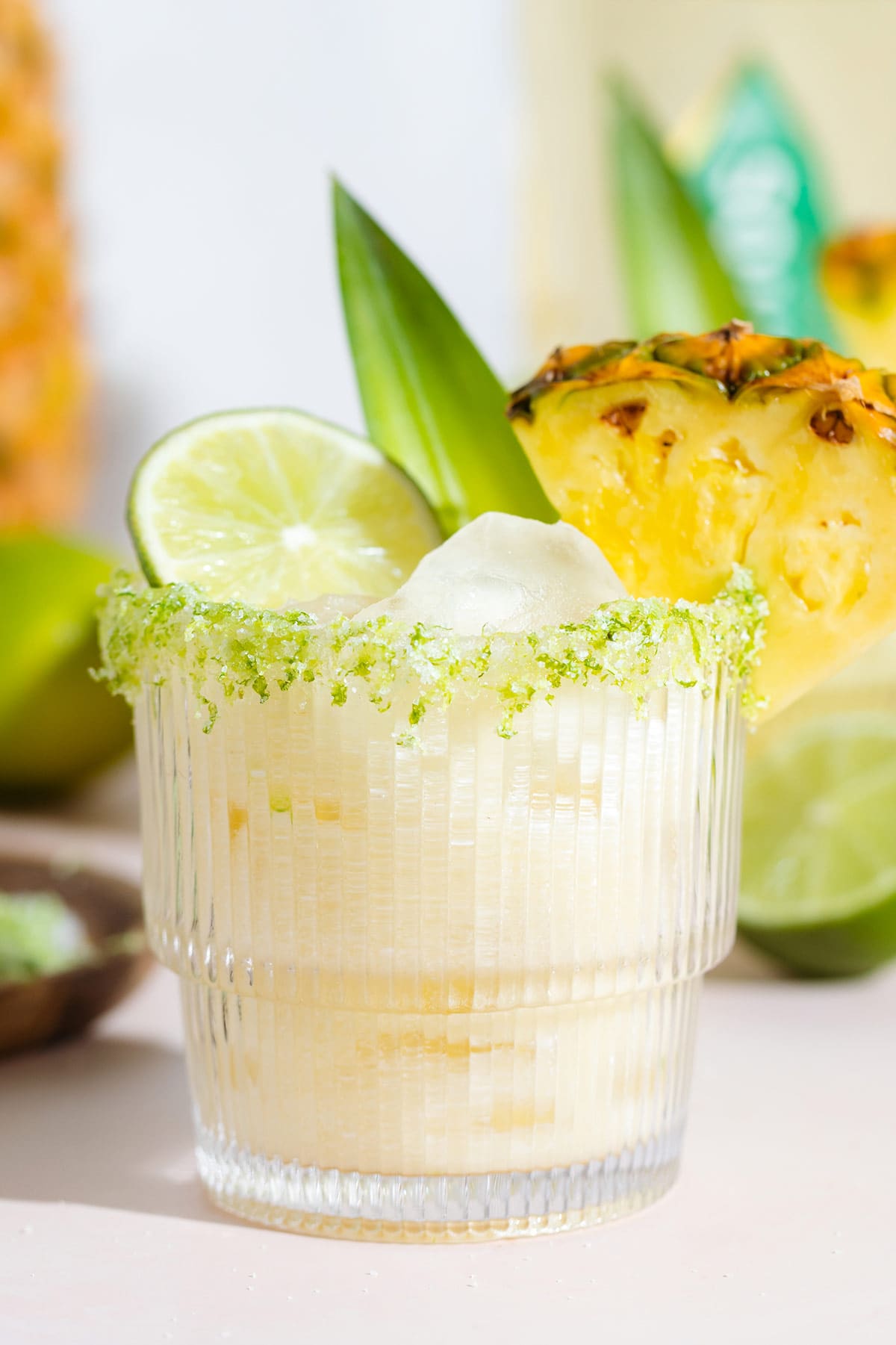 Pineapple coconut margarita in a short glass with salt and lime zest on the rim garnished with a slice of pineapple, a pineapple leaf, and lime.