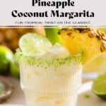 Pineapple coconut margarita in a short glass with salt and lime zest on the rim garnished with a slice of pineapple, a pineapple leaf, and lime.