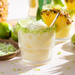 Pineapple coconut margarita in a short glass with salt and lime zest on the rim garnished with a slice of pineapple and lime with a short gold straw.