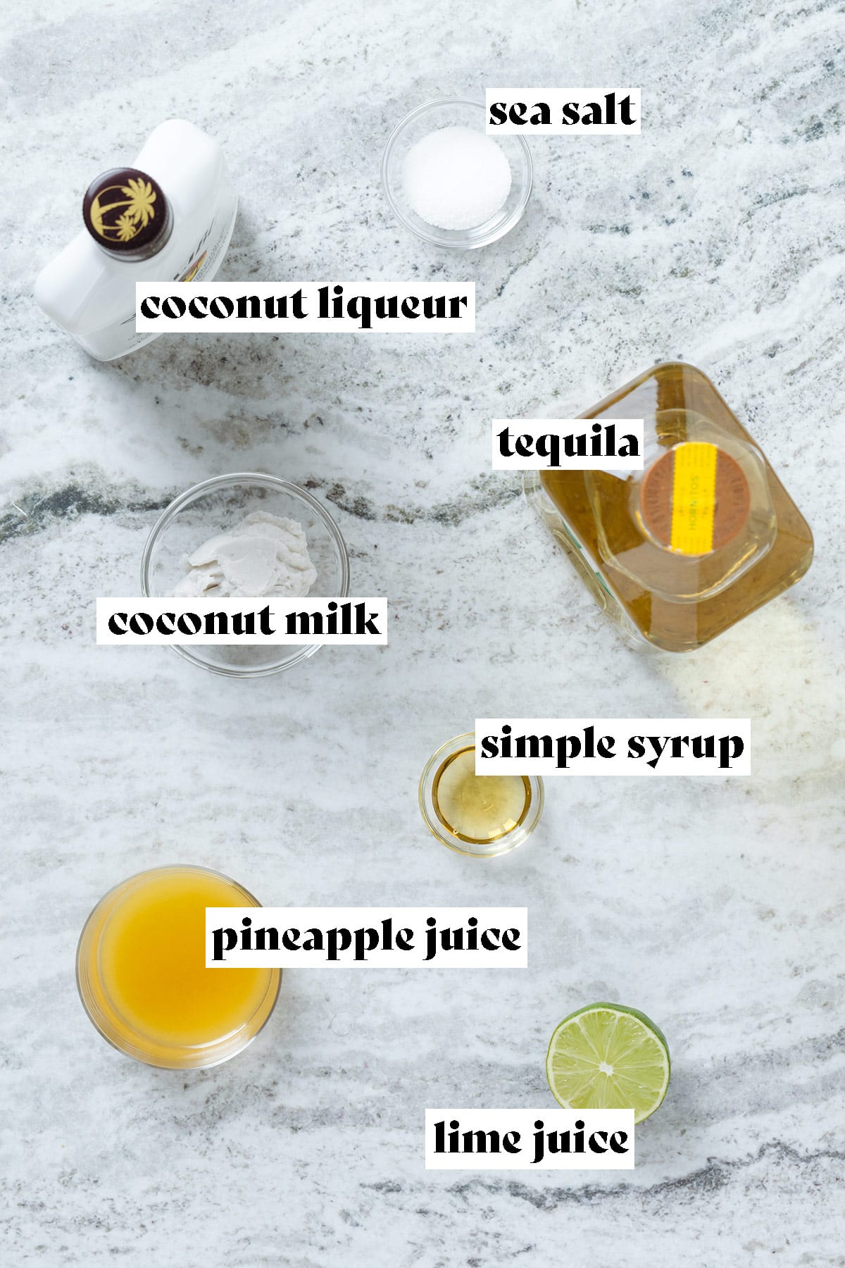 Ingredients like tequila, coconut rum, coconut cream, and pineapple juice laid out on a stone background.