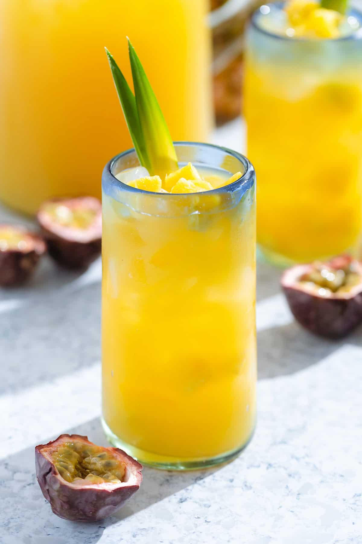 A bright yellow drink in a tall glass with a blue rim garnished with two pineapple leaves and diced pineapple.