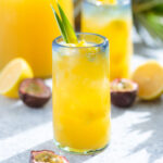 A bright yellow drink in a tall glass with a blue rim garnished with two pineapple leaves and diced pineapple.