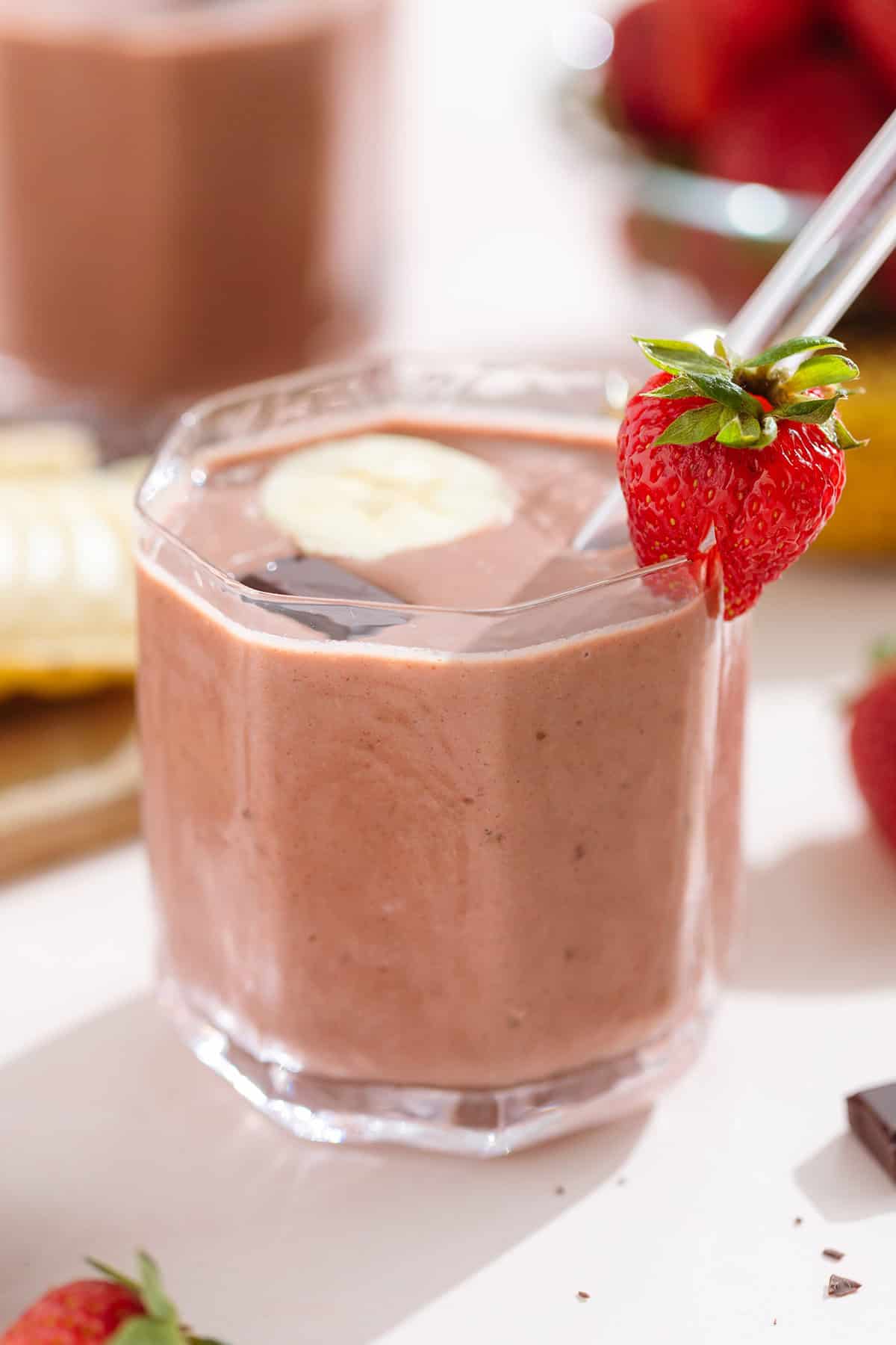 Strawberry smoothie in a short glass garnished with a strawberry, a banana slice, and a square of chocolate.