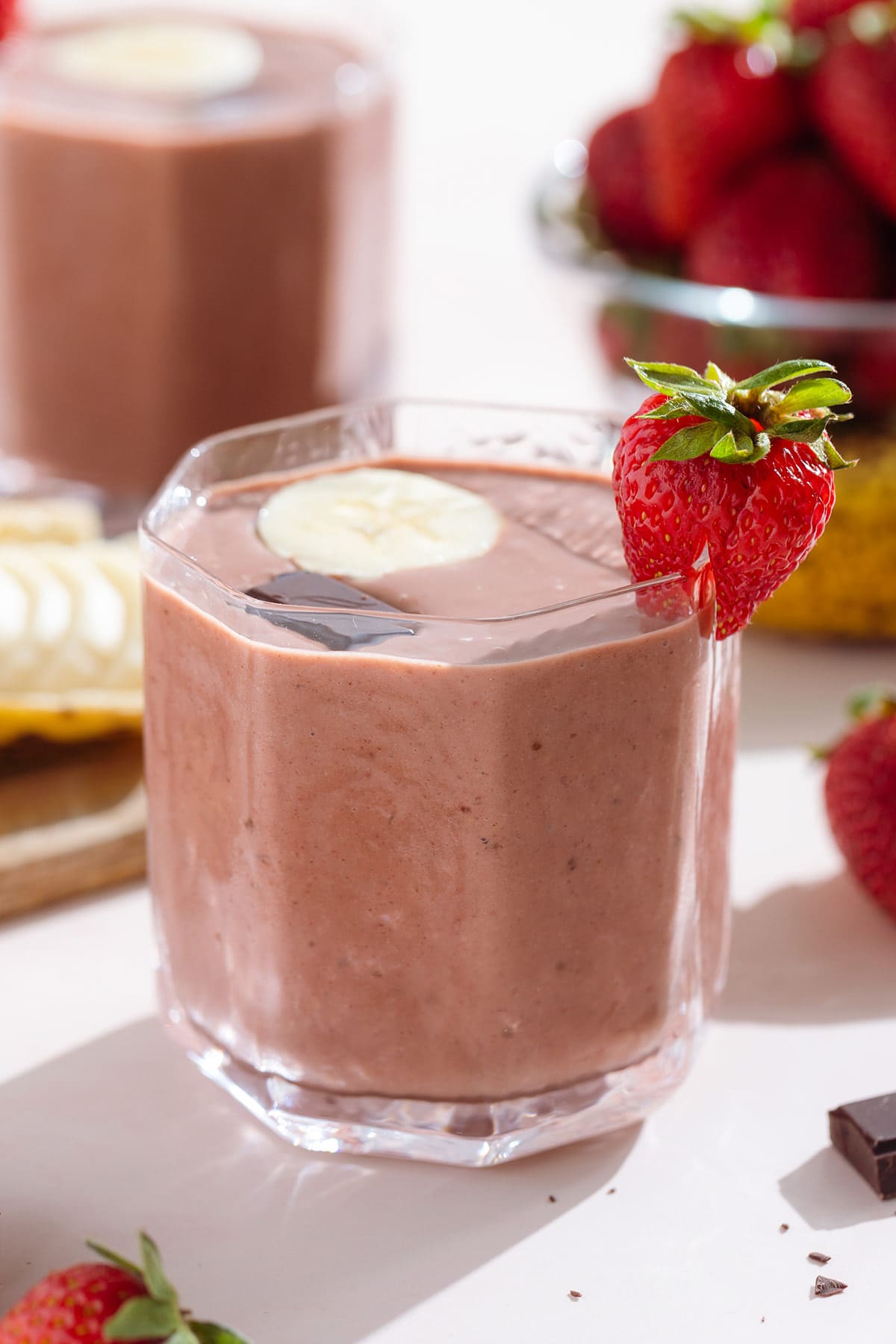 Strawberry smoothie in a short glass garnished with a strawberry, a banana slice, and a square of chocolate.