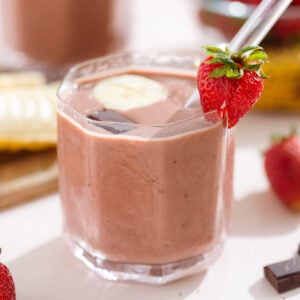 Strawberry smoothie in a short glass garnished with a strawberry, a banana slice, and a square of chocolate.
