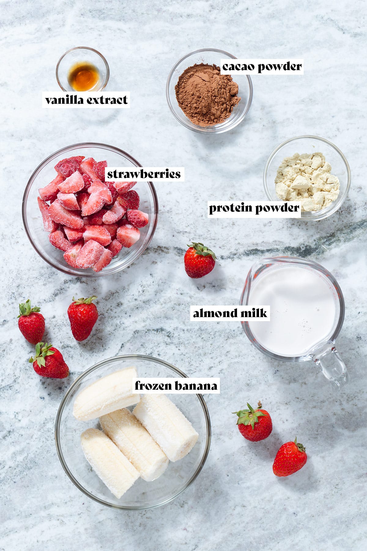 Strawberries, bananas, milk, cacao, and protein powder laid out on a grey stone background.