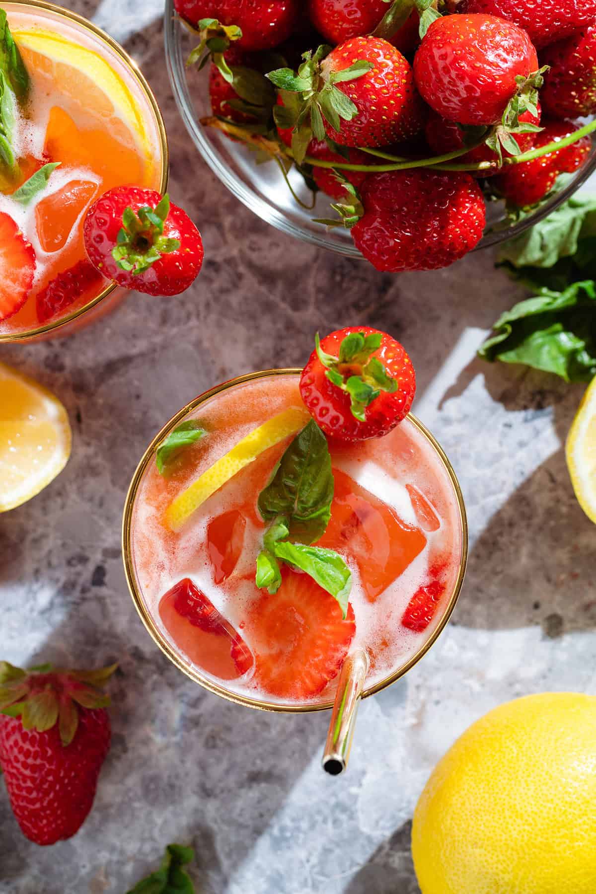 Bright red strawberry basil lemonade in a short glass with a gold rim garnished with a strawberry and fresh basil with a glass straw.