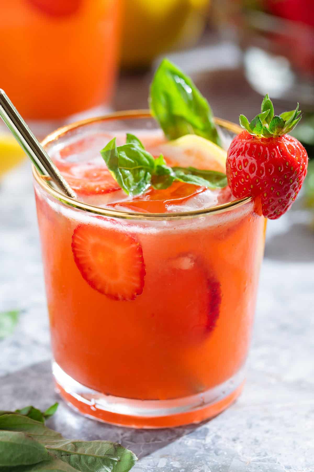 Bright red strawberry basil lemonade in a short glass with a gold rim garnished with a strawberry and fresh basil with a glass straw.