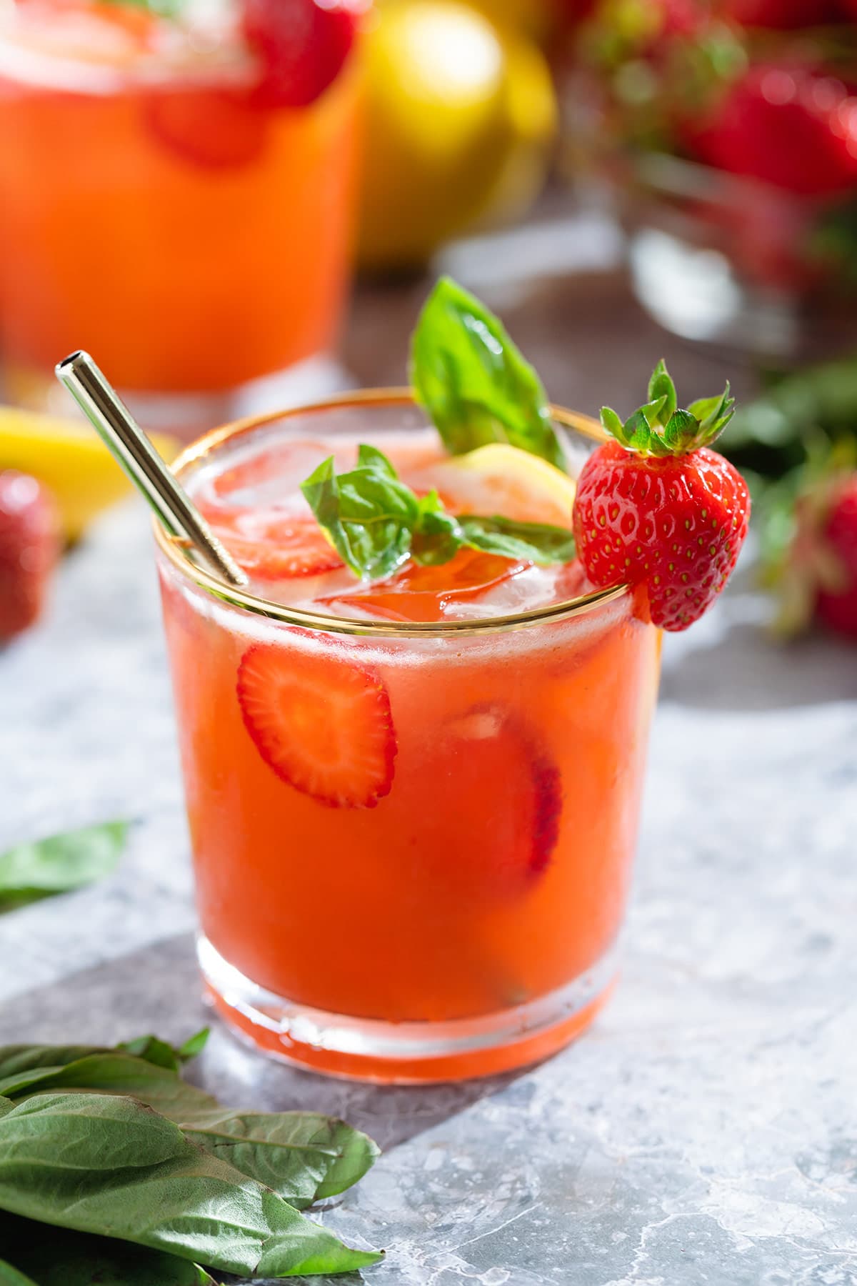 Bright red strawberry basil lemonade in a short glass with a gold rim garnished with a strawberry and fresh basil with a glass straw.