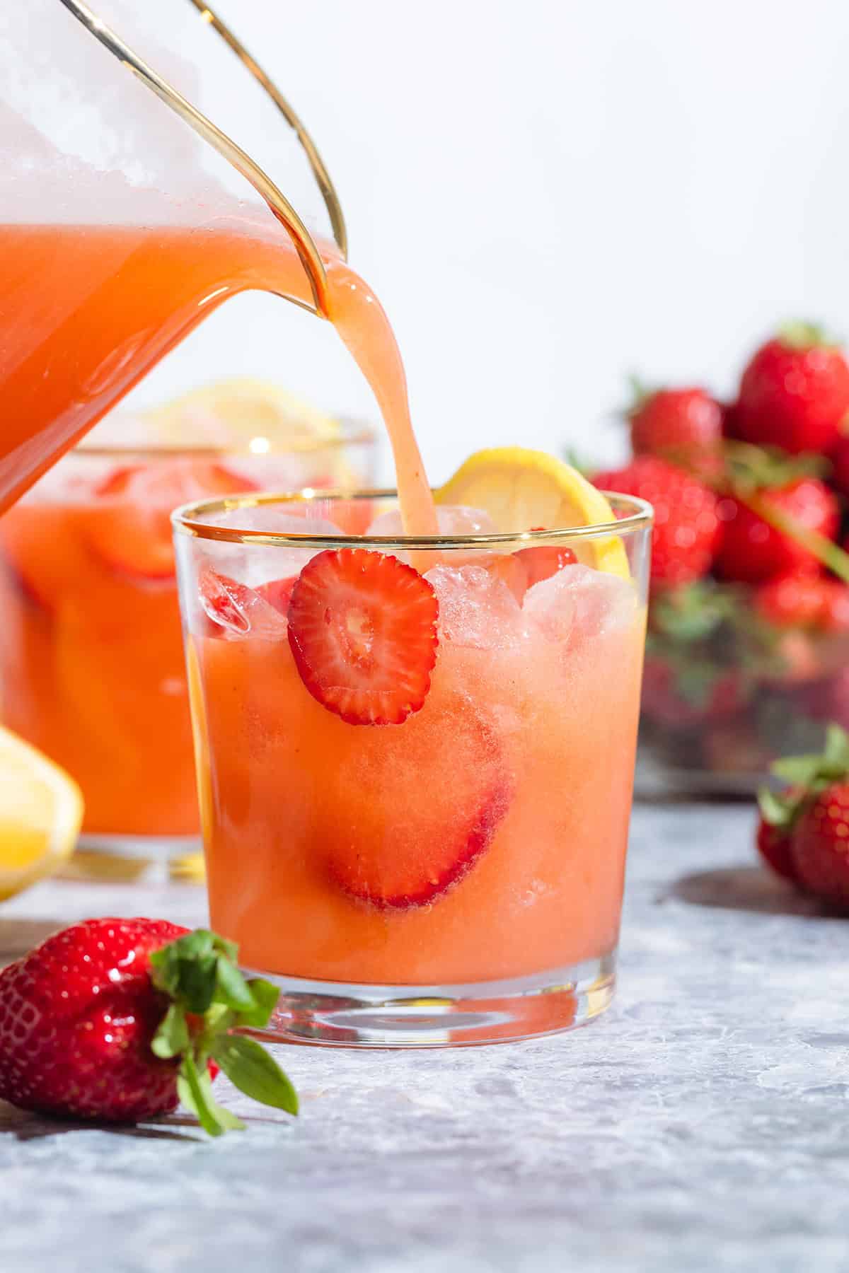 Bright red strawberry basil lemonade being poured into a short glass with a gold rim garnished with a slice of lemon.