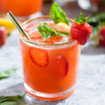 Bright red strawberry basil lemonade in a short glass with a gold rim garnished with a strawberry and fresh basil with a glass straw.