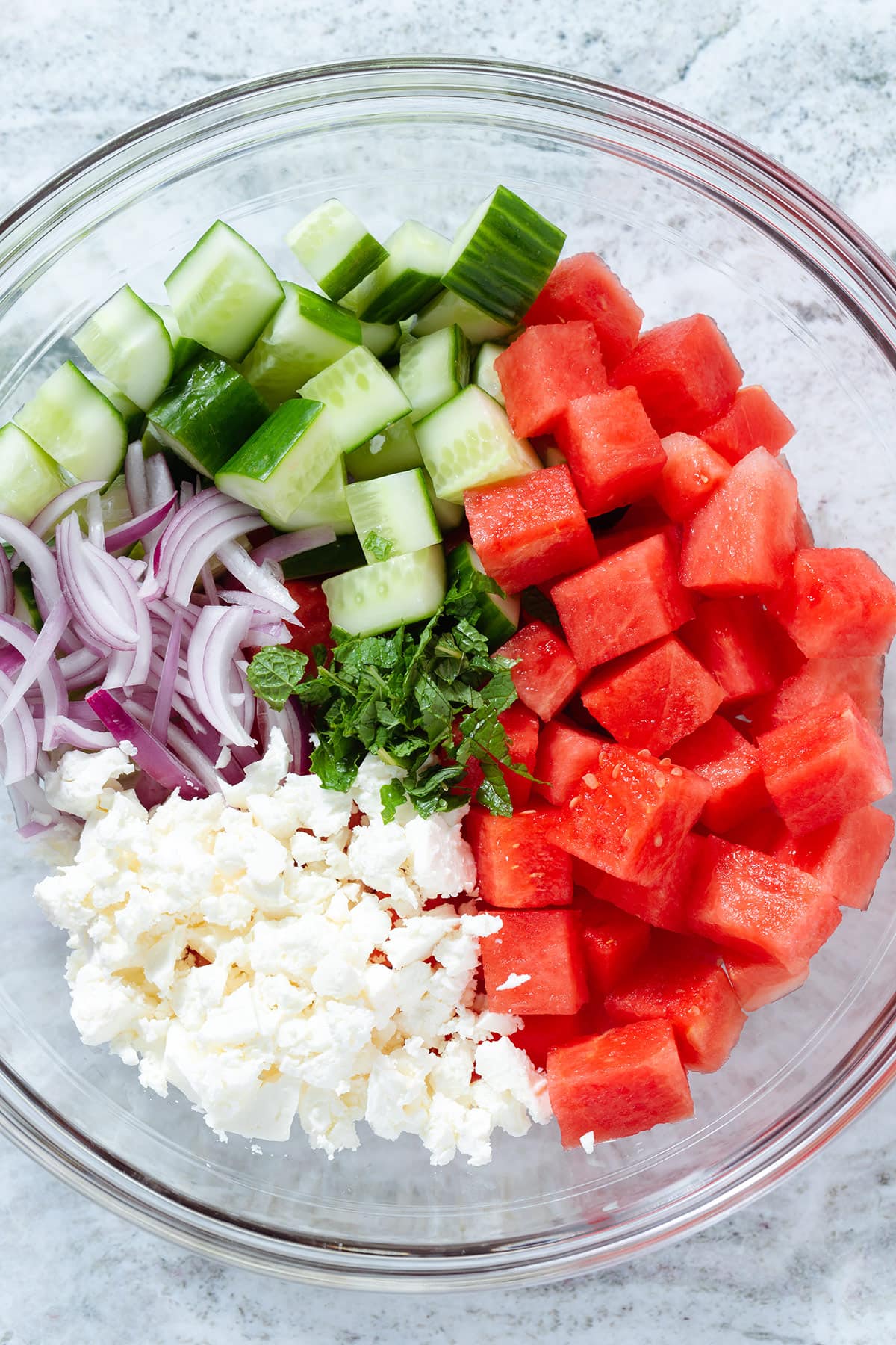 Diced cucumber, watermelon, feta, sliced red onion, and mint in a large glass bowl.