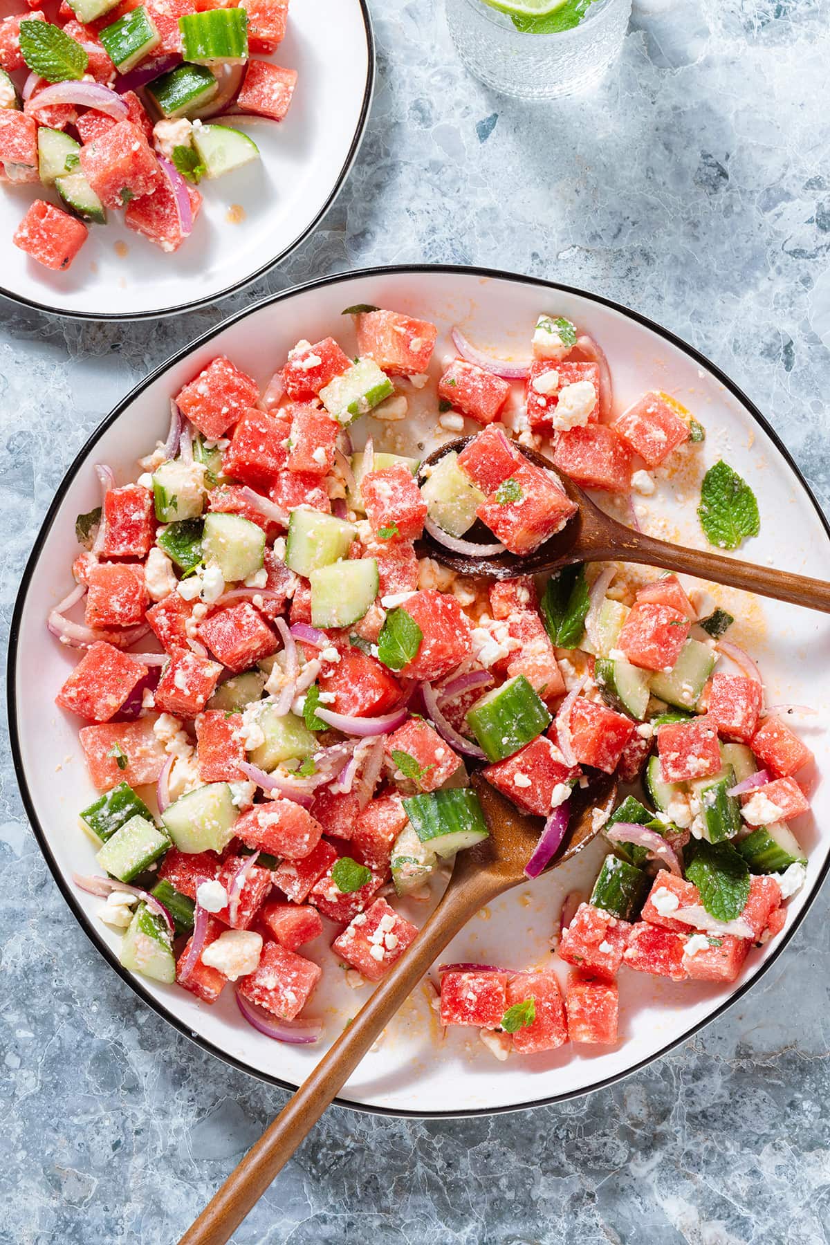 Watermelon salad on a large white serving platter with a black rim on a stone background with two wooden serving spoons inserted in.