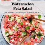 Watermelon salad on a large white serving platter with a black rim on a stone background with two wooden serving spoons inserted in.