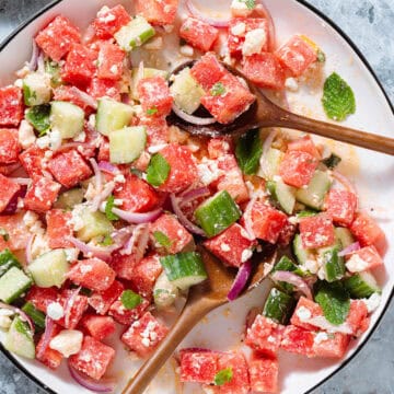 Watermelon salad on a large white serving platter with a black rim on a stone background with two wooden serving spoons inserted in.