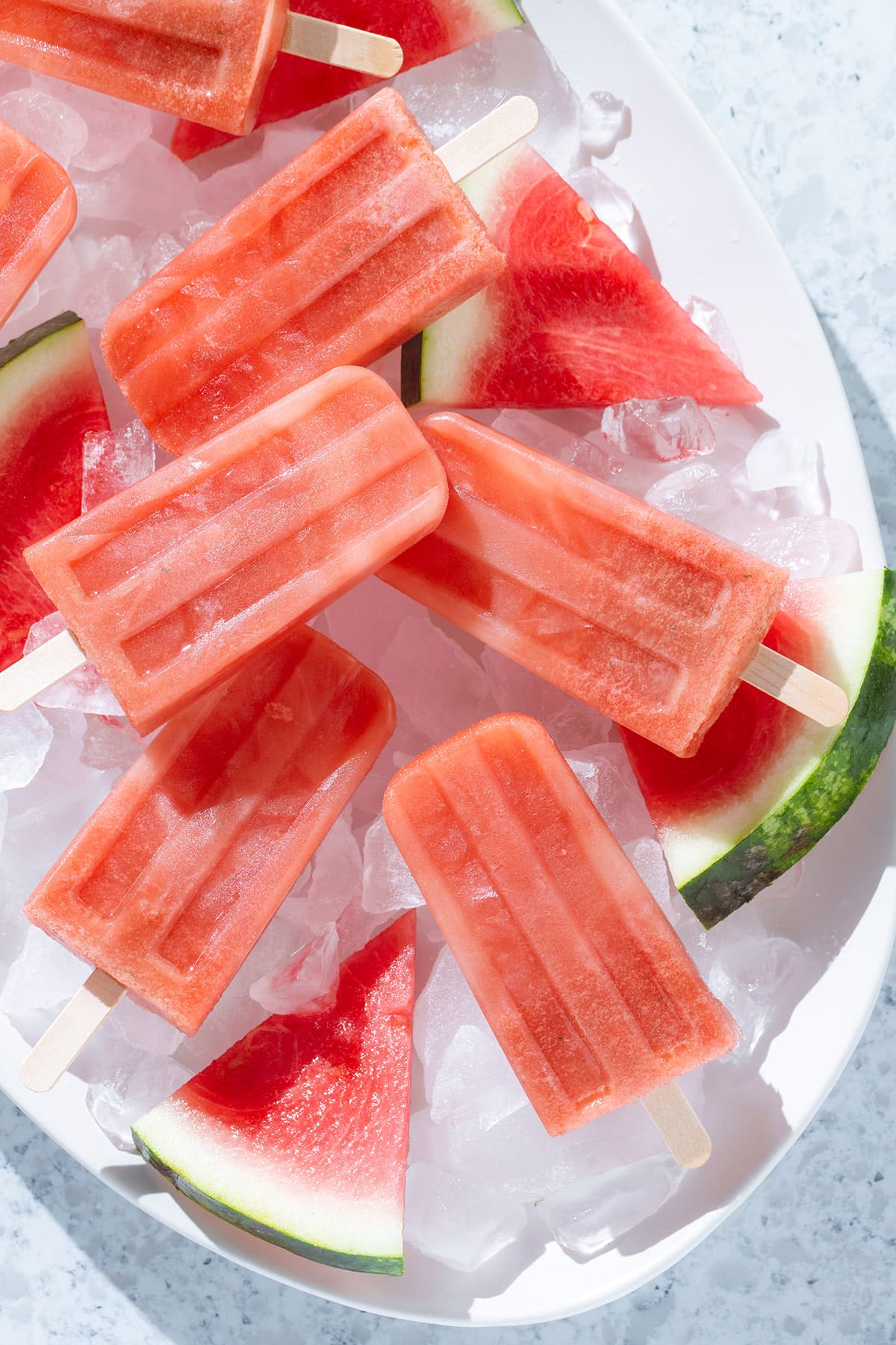 Watermelon popsicles on a large white serving platter with ice and slices of watermelon.