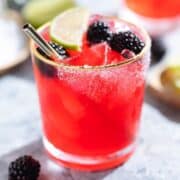 Bright purple red blackberry margarita in a glass with a gold rim garnished with blackberries and lime slices and salt on the rim on a grey stone background.