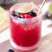 Bright pink purple margarita in a short glass garnished with blueberries and a lime slice with salt on the rim and a gold straw.