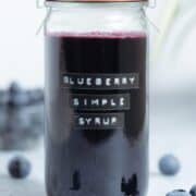 A glass jar with dark purple blueberry syrup with fresh blueberries around the jar and a green plant in the background.