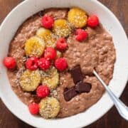Chocolate oatmeal in a beige bowl topped with fresh raspberries, caramelized banana slices and chocolate squares.