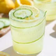 A short glass with light green cucumber lemonade over ice garnished with a slice of lemon and cucumber with more lemonade in the background.