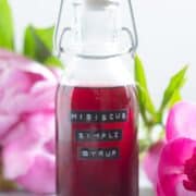 A small glass bottle with bright red hibiscus syrup with peonies in the background.