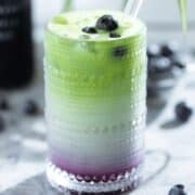 Iced matcha latte in a tall glass with a layer of blueberry syrup on the bottom of the glass garnished with fresh blueberries with a glass straw and a bottle of blueberry syrup in the background.