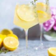 A tall white wine glass with a lemon spritzer garnished with lemon slices and fresh thyme with more lemons around the glass.