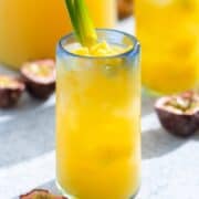 A bright yellow drink in a tall glass with a blue rim garnished with two pineapple leaves and diced pineapple.