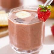 Strawberry smoothie in a short glass garnished with a strawberry, a banana slice, and a square of chocolate.
