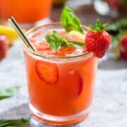 Bright red strawberry basil lemonade in a short glass with a gold rim garnished with a strawberry and fresh basil with a glass straw.