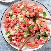 Watermelon salad on a large white serving platter with a black rim on a stone background with two wooden serving spoons inserted in.