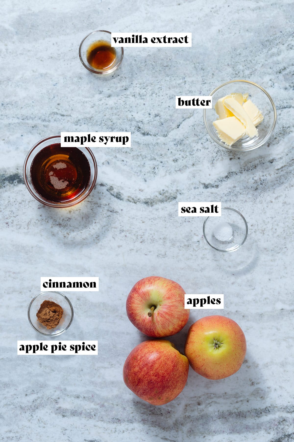 Ingredients like apples, butter, and maple syrup laid out on a grey stone background.