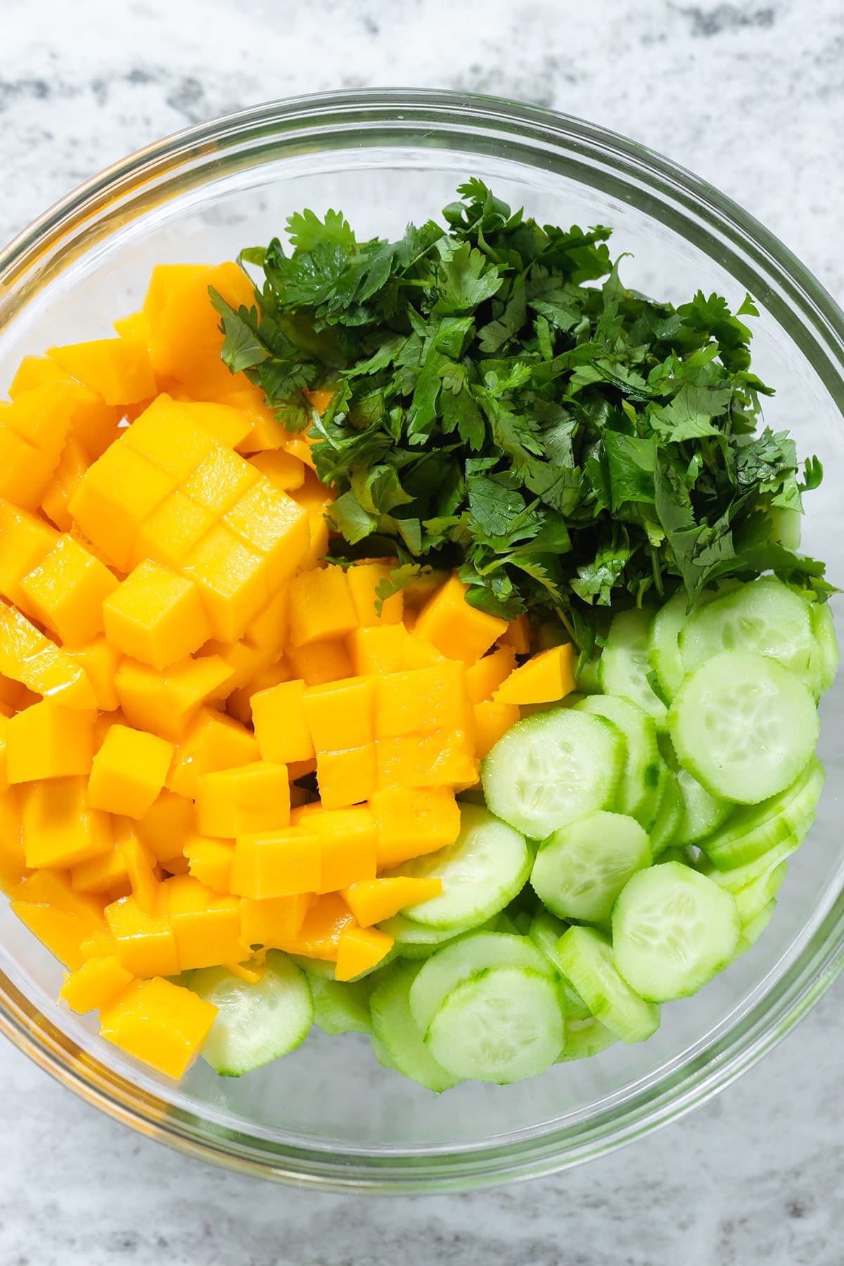 Diced mango, sliced cucumber, and chopped cilantro in a glass bowl on a grey stone background.