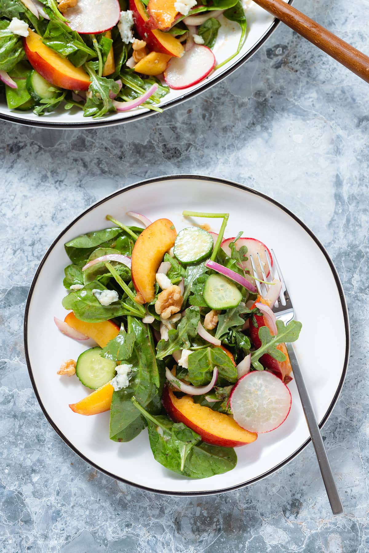 Nectarine salad with mixed greens, red onion, cucumbers, radishes, and blue cheese on a small white salad plate with a black rim and a silver fork on the right side.