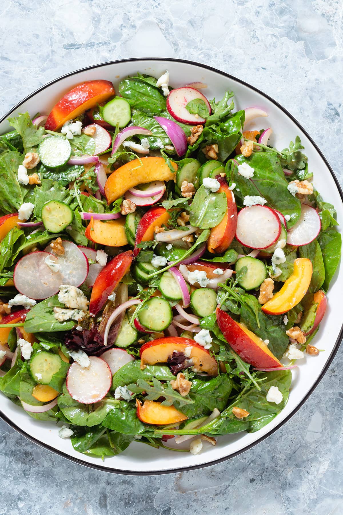 Nectarine salad with mixed greens and radishes on a large white serving platter with a black rim on a grey stone background.