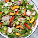 Nectarine salad with mixed greens and radishes on a large white serving platter with a black rim on a grey stone background.