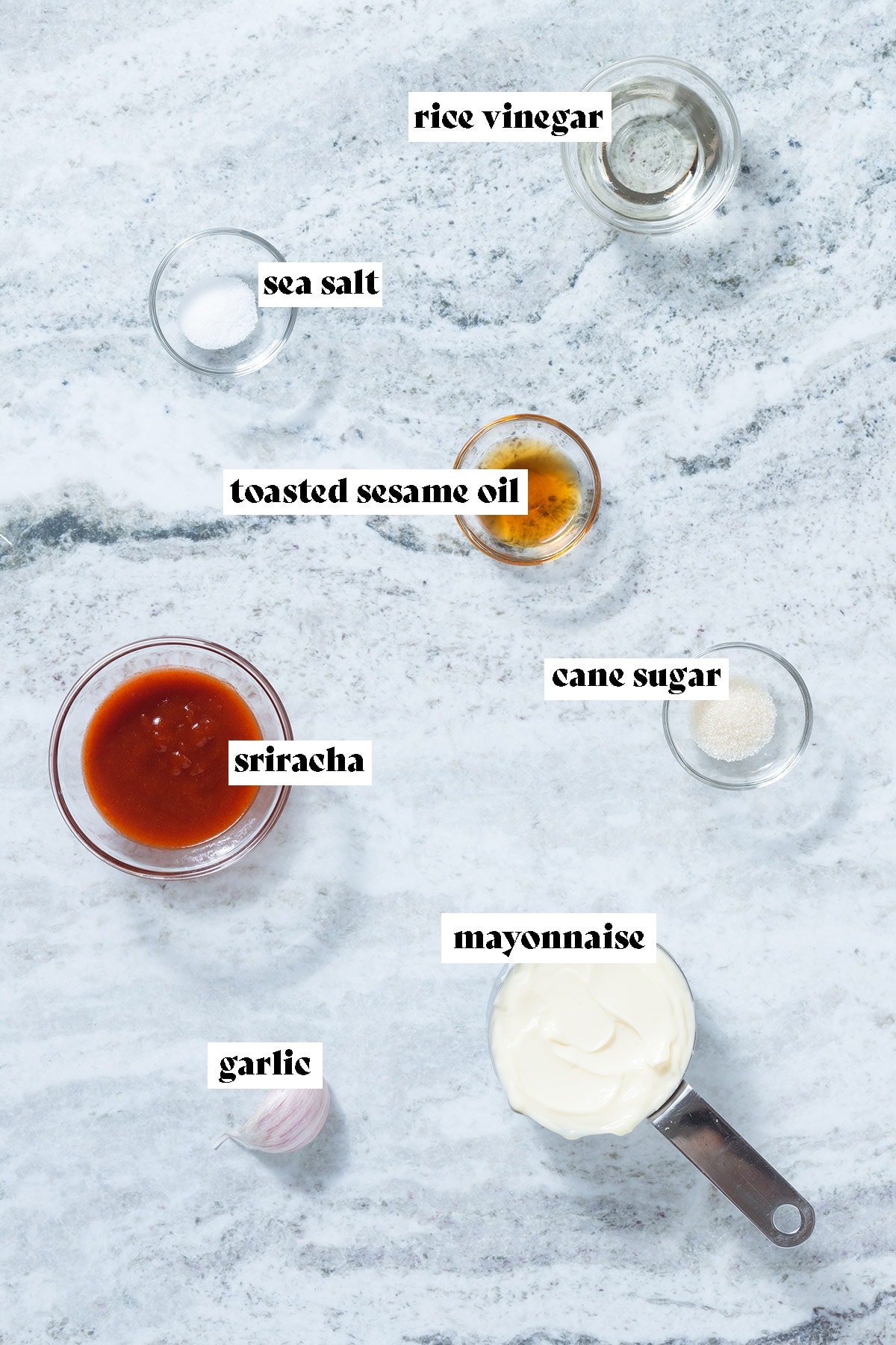 Sriracha, mayonnaise, sesame oil, and other ingredients laid out on a grey stone background.