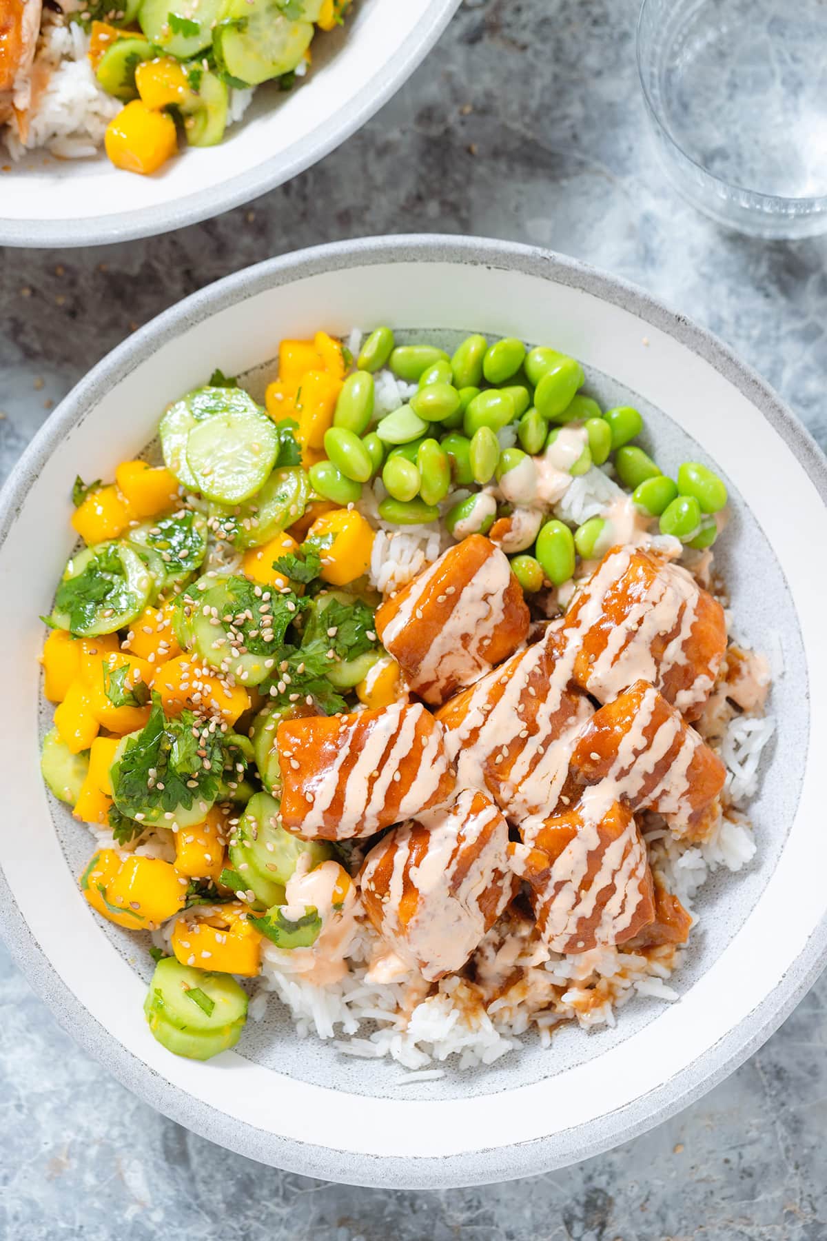 Diced teriyaki salmon with mango salad, edamame, and rice drizzled with spicy mayo in a grey bowl on a grey background.