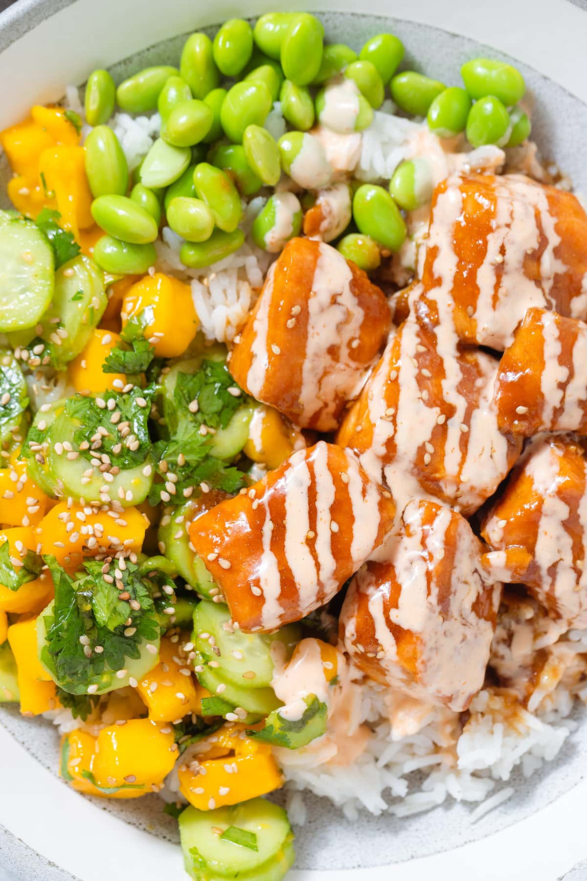 A close up of diced teriyaki salmon with mango salad, edamame, and rice drizzled with spicy mayo in a grey bowl.