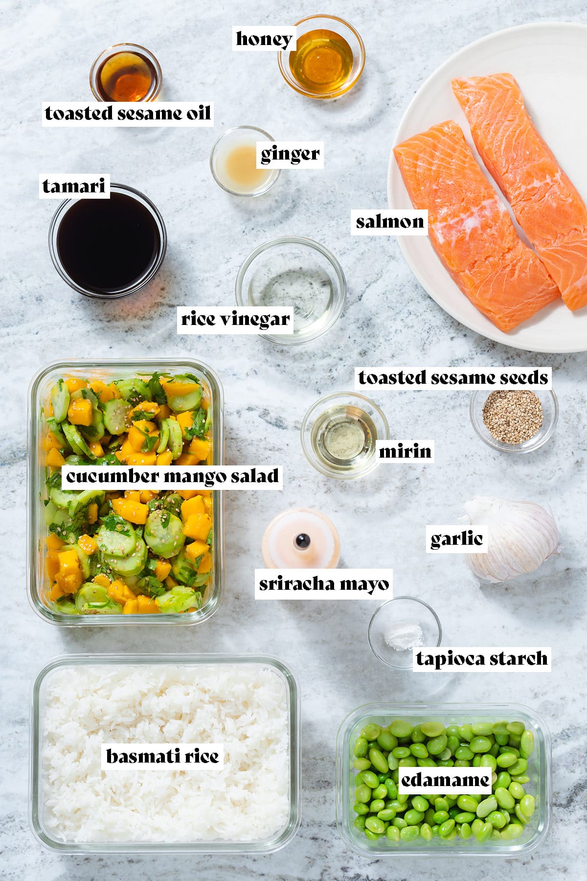 Ingredients like raw salmon, mango salad, rice, edamame, and spices laid out on a grey background.